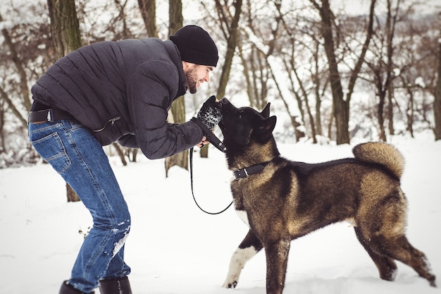 Un hombre con una chaqueta y un gorro de punto camina por un bosque nevado con un perro Akita americano