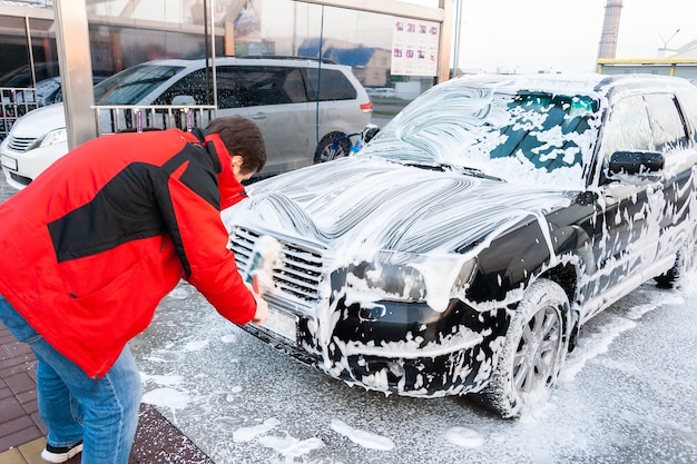 Un hombre con una chaqueta roja limpia un auto negro cubierto de espuma con un cepillo en un autoservicio de lavado de autos. Vista frontal