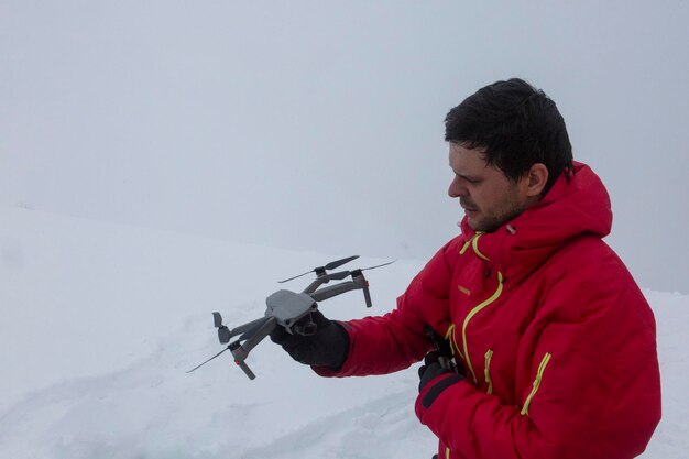 Un hombre con una chaqueta roja sostiene un dron frente a la nieve.