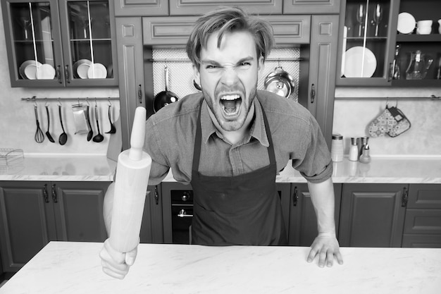 Hombre chef en delantal rojo gritar con ira con rodillo en la mesa en la cocina Cocinar hornear cocina culinaria Concepto de comida casera