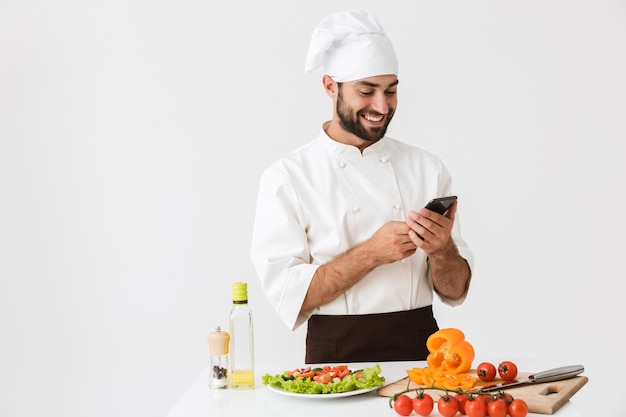 Hombre chef satisfecho en uniforme sonriendo y sosteniendo el teléfono inteligente mientras cocina ensalada de verduras aislado sobre la pared blanca