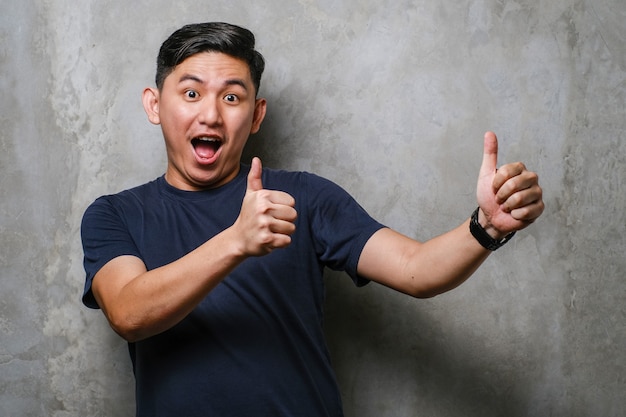 Hombre chino guapo joven con camiseta casual sobre fondo de muro de hormigón aprobando hacer un gesto positivo con la mano, pulgar hacia arriba sonriendo y feliz por el éxito. Gesto de ganador.