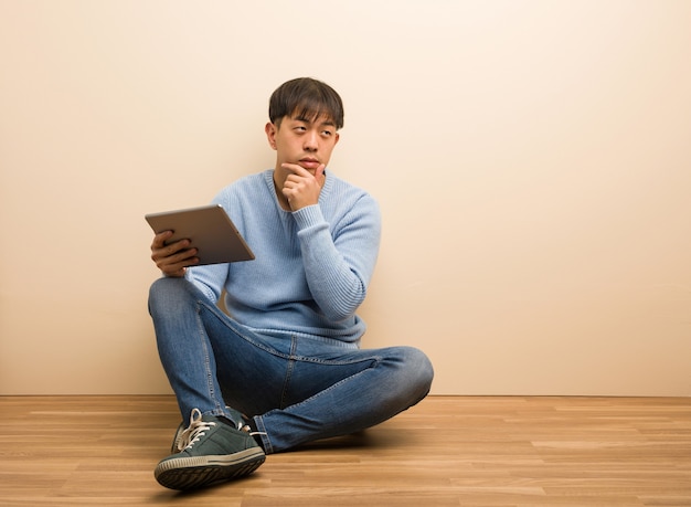 Hombre chino joven sentado usando su tableta dudando y confundido