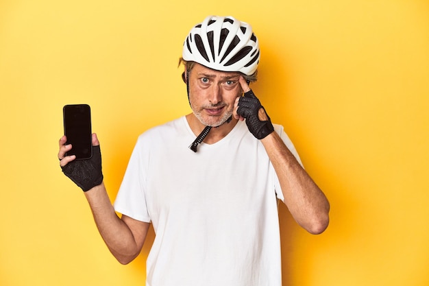 Hombre ciclista mostrando teléfono sobre fondo amarillo mostrando un gesto de decepción con el dedo índice