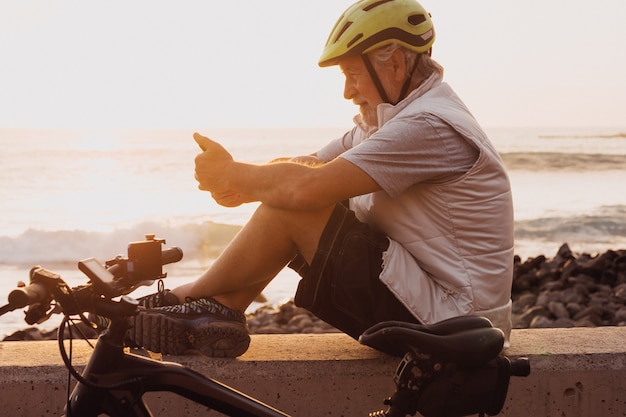 Hombre ciclista con teléfono móvil sentado junto al mar al atardecer, cierra su bicicleta. Personas jubiladas que disfrutan de un estilo de vida saludable