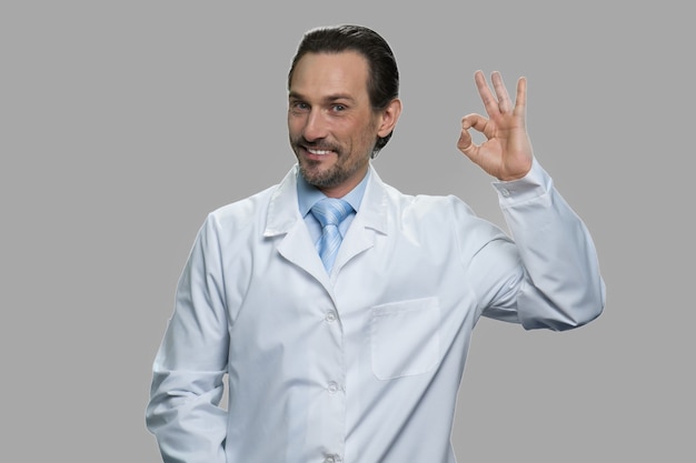 Hombre científico caucásico mostrando ok con los dedos. Retrato de hombre de mediana edad alegre en bata de laboratorio mirando a cámara sobre fondo gris.