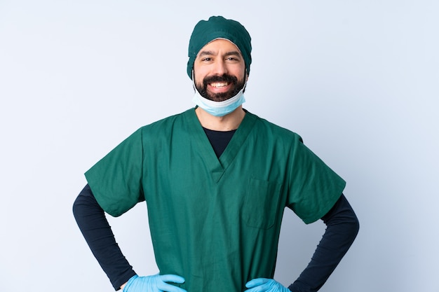 Hombre cirujano en uniforme verde sobre pared aislada posando con los brazos en la cadera y sonriendo