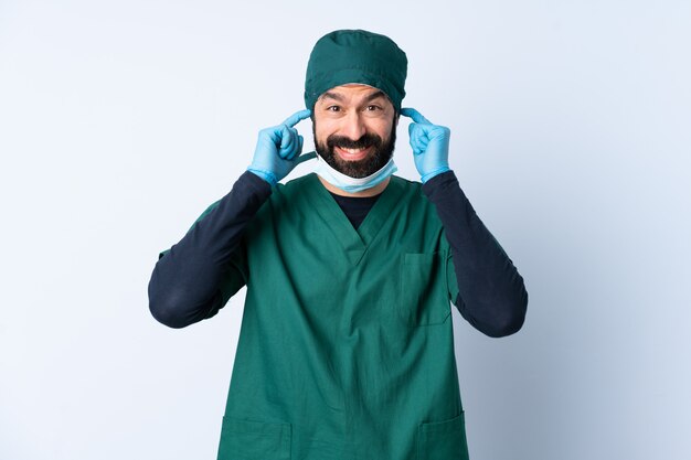 Hombre cirujano en uniforme verde sobre pared frustrado y cubriendo las orejas