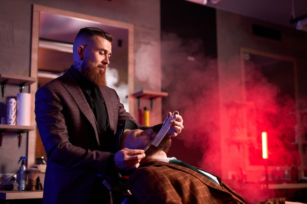 Hombre de cliente caucásico hipster visitando en peluquería afeitado barba por peluquero profesional