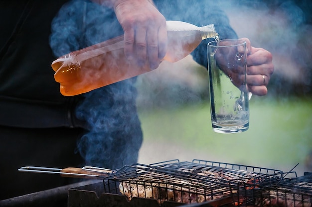 un hombre cocina carne a la parrilla y vierte cerveza fría y espumosa de una botella en un vaso en la naturaleza