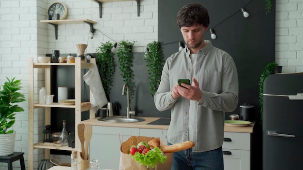 El hombre se para en la cocina cerca de una bolsa de papel llena de comida fresca y usa una aplicación de teléfono inteligente para entregarla en el supermercado.