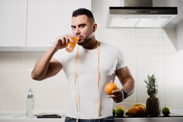 Hombre en la cocina sosteniendo una naranja y bebiendo jugo en la cocina. El concepto de un estilo de vida saludable.