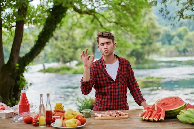 Hombre cocinando comida sabrosa en la parrilla de la barbacoa para una cena francesa al aire libre cerca del río en una hermosa noche de verano en la naturaleza