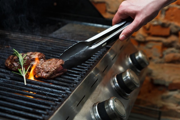 Hombre cocinando filetes de carne en parrilla profesional al aire libre. Mano masculina con pinzas girando bistecs.