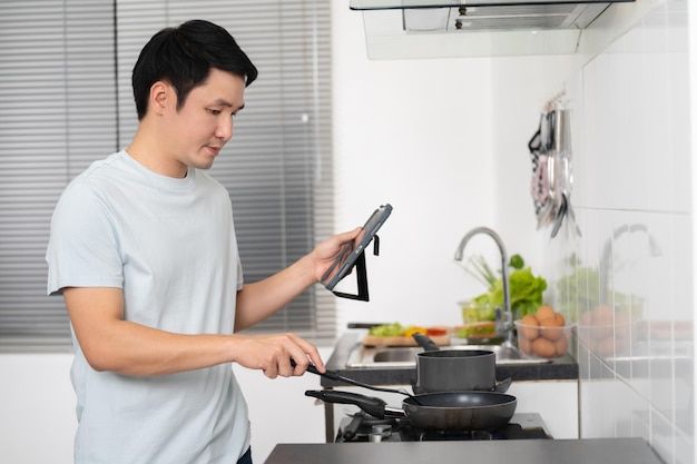 Hombre cocinando y preparando comida según una receta en una tableta en la cocina de casa