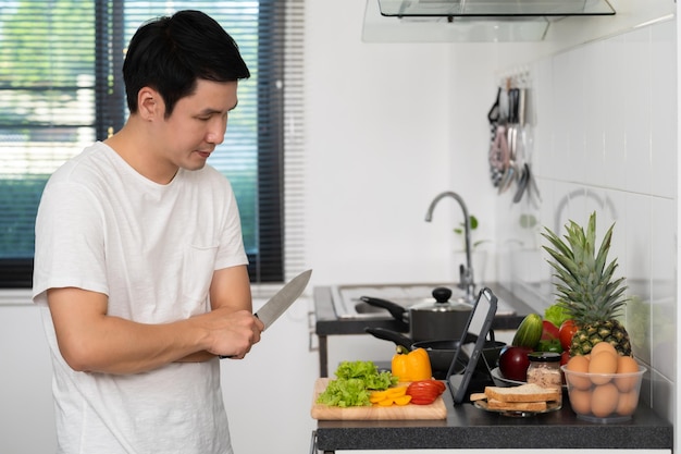 Hombre cocinando y preparando verduras según una receta en una tableta en la cocina de casa