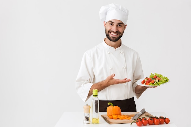 Hombre cocinero positivo en uniforme sonriendo y sosteniendo el plato con ensalada de verduras aislado sobre la pared blanca