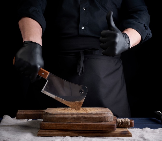 Hombre cocinero en uniforme negro y guantes de látex negro sostiene un cuchillo de carne afilado grande sobre una tabla de cortar