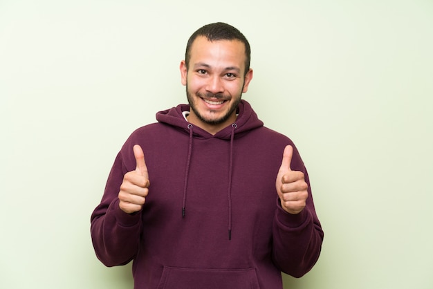 Hombre colombiano con sudadera sobre muro verde dando un gesto con los pulgares arriba.