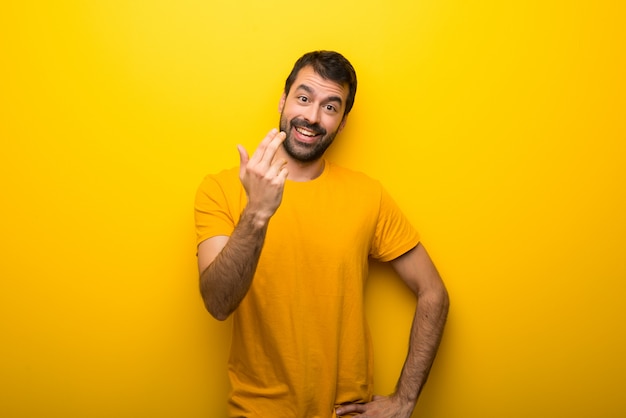 Hombre en el color amarillo vibrante aislado que invita para venir con la mano. Feliz de que hayas venido