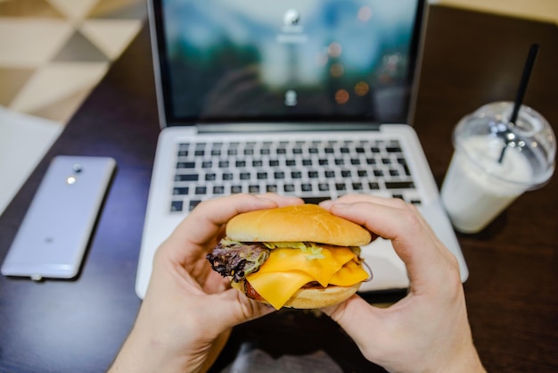 El hombre come hamburguesas en la cafetería y trabaja en la computadora portátil