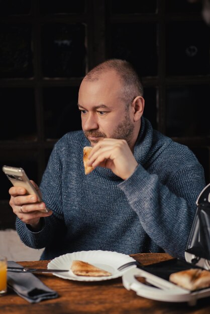 Un hombre come un sándwich y mira el teléfono en casa Un hombre ordinario de mediana edad Comiendo con un teléfono en la mano