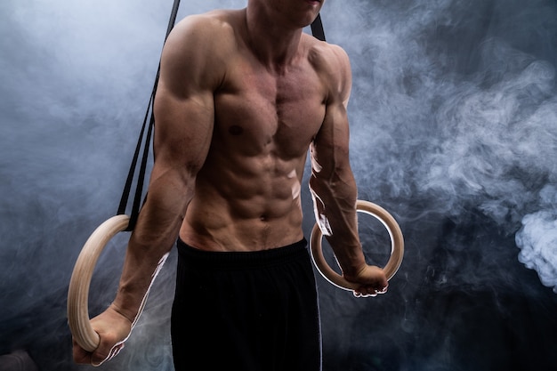 Hombre de complexión muscular haciendo crossfit en anillos de gimnasia interior sobre fondo negro, ahumado