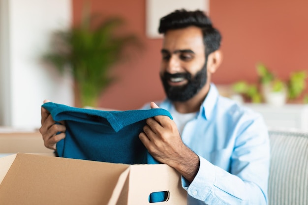 Hombre comprador indio contento desempaquetando caja con ropa en el interior