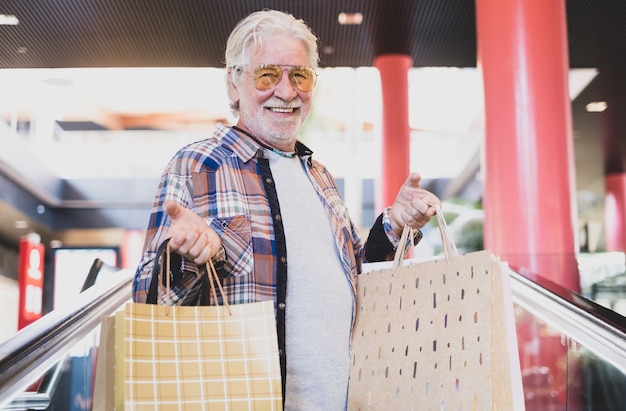 Hombre de compras. Hombre barbudo senior feliz con bolsas de compras en la escalera mecánica en el centro comercial mirando a la cámara sonriendo, concepto de consumismo