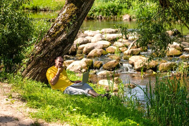 Hombre con computadora portátil y teléfono inteligente trabajando afuera en el prado junto al concepto de oficina al aire libre del río