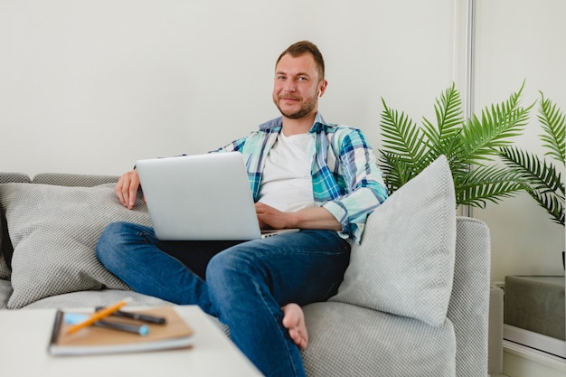 Hombre concentrado ocupado guapo en camisa sentado relajado en el sofá en casa en la mesa trabajando en línea en la computadora portátil desde casa