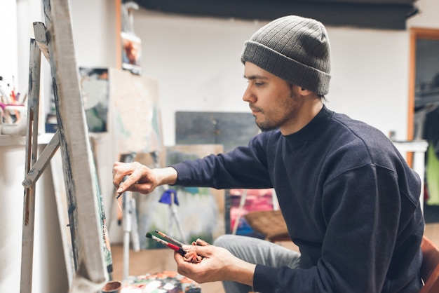 Hombre concentrado de pie en un estudio cerca del lienzo y pinta un cuadro