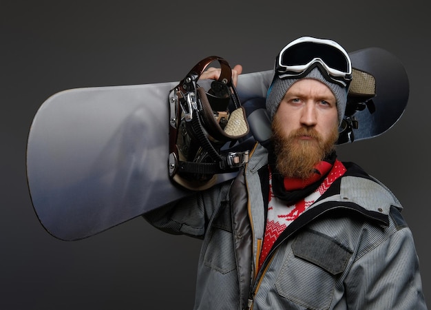 Foto hombre confiado con barba roja usando un equipo completo sosteniendo una tabla de snowboard en su hombro, mirando una cámara con una mirada seria