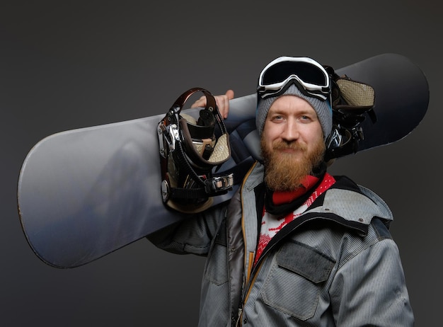 Hombre confiado con barba roja usando un equipo completo sosteniendo una tabla de snowboard en su hombro, sonriendo y mirando una cámara, aislado en un fondo oscuro.