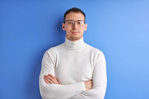 Hombre confiado en gafas posando mirando a la cámara con los brazos cruzados, vestido con camisa casual blanca, aislado sobre fondo azul de estudio