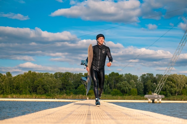 Hombre confiado en traje de neopreno con wakeboard caminando sobre un puente flotante después del entrenamiento