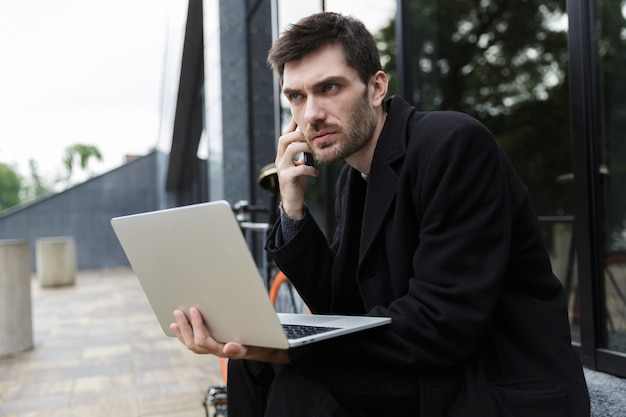 Hombre confiado vestido con abrigo sentado en la calle, usando la computadora portátil, hablando por teléfono móvil