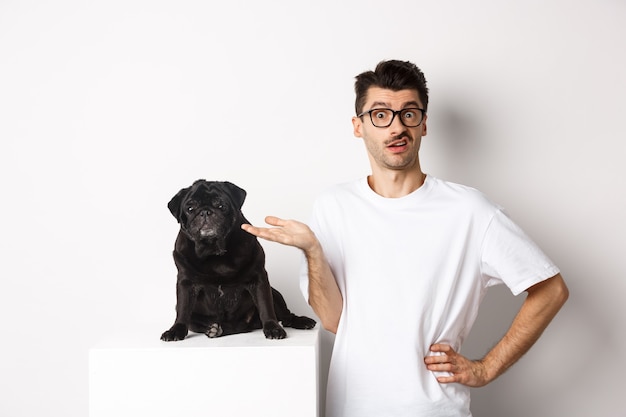 Foto hombre confundido al ver algo extraño, mirando a la cámara desconcertado, de pie cerca de la linda mascota pug negro contra blanco