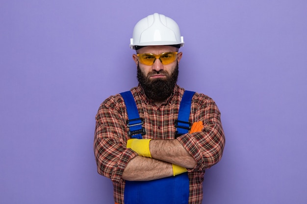 Hombre constructor barbudo en uniforme de construcción y casco de seguridad con guantes de goma mirando con el ceño fruncido serio con los brazos cruzados