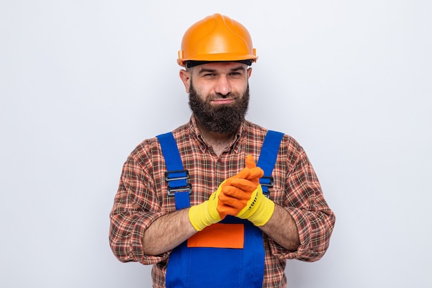 Hombre constructor barbudo en uniforme de construcción y casco de seguridad con guantes de goma mirando feliz y complacido frotándose las manos