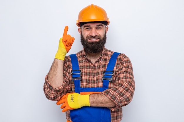 Hombre constructor barbudo en uniforme de construcción y casco de seguridad con guantes de goma mirando sonriendo alegremente mostrando el dedo índice