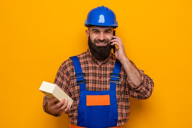 Hombre constructor barbudo en uniforme de construcción y casco de seguridad sosteniendo ladrillo sonriendo alegremente mientras habla por teléfono móvil de pie sobre fondo naranja