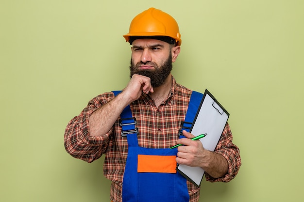 Hombre constructor barbudo en uniforme de construcción y casco de seguridad sosteniendo portapapeles con lápiz mirando a un lado desconcertado de pie sobre fondo verde