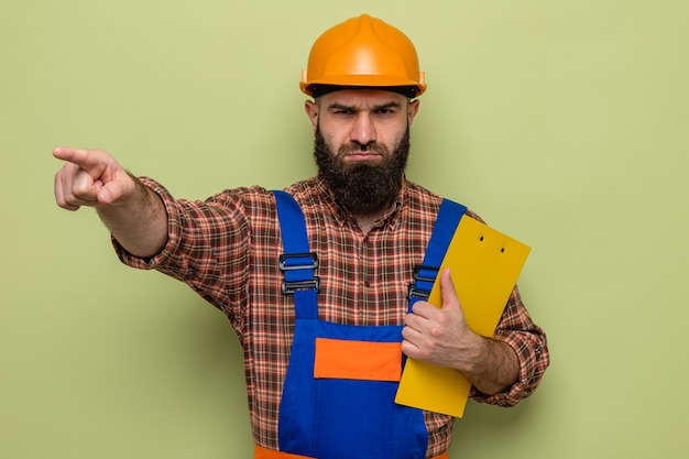 Hombre constructor barbudo en uniforme de construcción y casco de seguridad sosteniendo portapapeles mirando a cámara con el ceño fruncido apuntando con el dedo índice a algo de pie sobre fondo verde