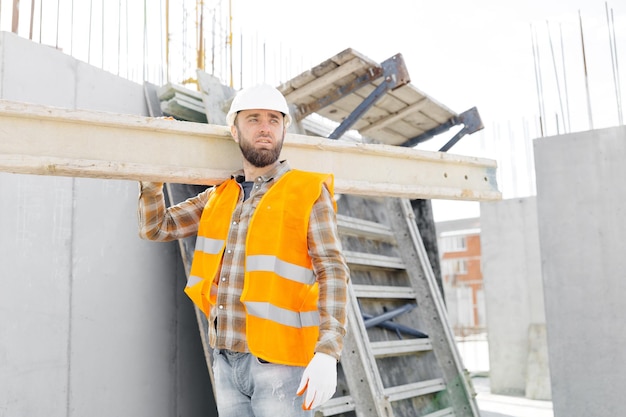 Hombre constructor con casco y chaleco cargando madera en el sitio de construcción en su jornada laboral