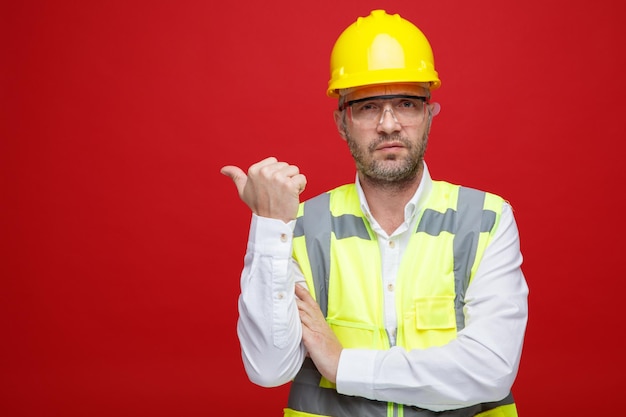 Hombre constructor en uniforme de construcción y casco de seguridad con gafas de seguridad mirando a la cámara con cara seria apuntando con el pulgar hacia el lado de pie sobre fondo rosa