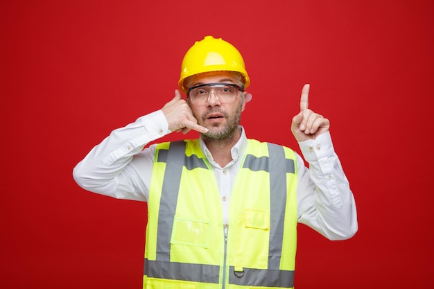 Hombre constructor en uniforme de construcción y casco de seguridad con gafas de seguridad mirando a la cámara haciendo un gesto de llamarme apuntando con el dedo índice hacia arriba de pie sobre fondo rojo