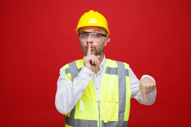 Hombre constructor en uniforme de construcción y casco de seguridad con gafas de seguridad mirando a la cámara haciendo gesto de silencio con el dedo en los labios apuntando con el dedo índice hacia abajo de pie sobre fondo rojo