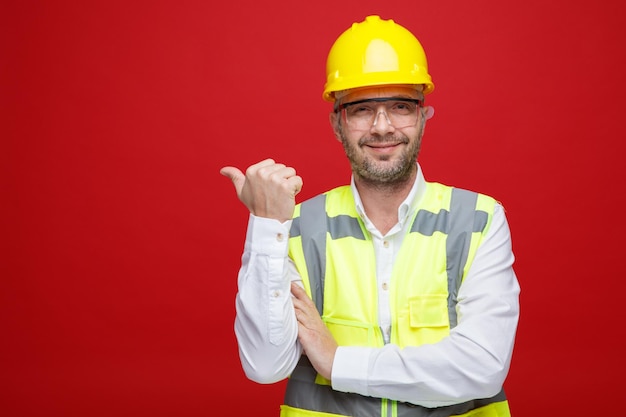 Hombre constructor en uniforme de construcción y casco de seguridad con gafas de seguridad mirando a la cámara con una sonrisa en la cara apuntando con el pulgar hacia el lado de pie sobre fondo rosa