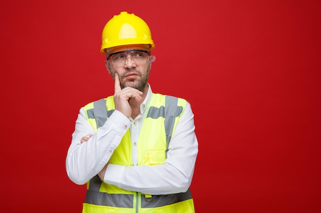 Hombre constructor en uniforme de construcción y casco de seguridad con gafas de seguridad mirando a un lado con expresión pensativa pensando de pie sobre fondo rosa